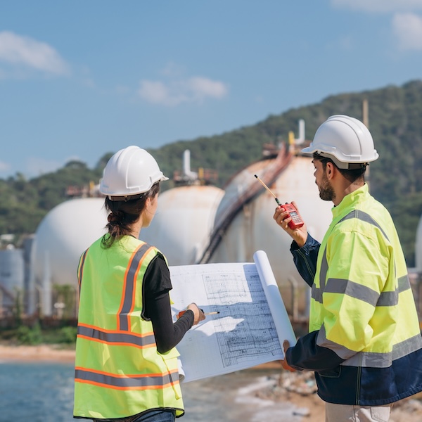 Two engineers in safety gear with blueprints discussing work in front of industrial storage tanks on a clear day.